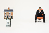Foto: House on top of roll of bills with pensive businessman on chair representing expensive real estate House on top of roll of bills with pensive businessman on chair representing expensive real estate