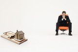 Foto: Model home on mouse trap with worried businessman sitting on chair representing increasing real estate rates Model home on mouse trap with worried businessman sitting on chair representing increasing real estate rates