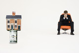 Foto: House on top of roll of bills with worried businessman on chair representing expensive real estate House on top of roll of bills with worried businessman on chair representing expensive real estate