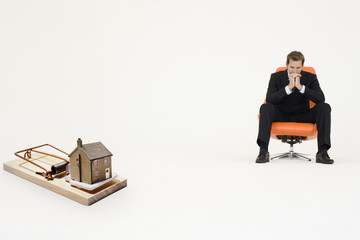 Photo: Model home on mouse trap with worried businessman sitting on chair representing increasing real estate rates Model home on mouse trap with worried businessman sitting on chair representing increasing real estate rates