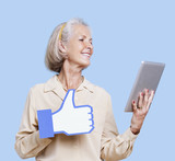 Photo : Senior woman with tablet PC holding fake like button against blue background Senior woman with tablet PC holding fake like button against blue background