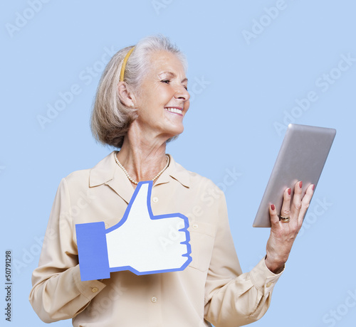 Photo: Senior woman with tablet PC holding fake like button against blue background Senior woman with tablet PC holding fake like button against blue background