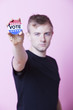 Photo: Portrait of a young man holding a VOTE badge up to the camera against pink background Portrait of a young man holding a VOTE badge up to the camera against pink background