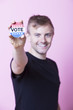 Photo: Portrait of a young man holding a VOTE badge up to the camera against pink background Portrait of a young man holding a VOTE badge up to the camera against pink background