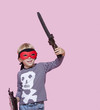 Photo: Happy young boy wearing eye mask holding sword and shield over pink background Happy young boy wearing eye mask holding sword and shield over pink background
