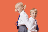 Photo: Happy young boy and girl in school uniform standing back to back over orange background Happy young boy and girl in school uniform standing back to back over orange background