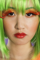 Photo: Close-up portrait of young woman wearing green wig and orange eyelashes over gray background Close-up portrait of young woman wearing green wig and orange eyelashes over gray background