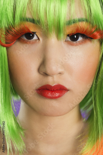 Photo: Close-up portrait of young woman wearing green wig and orange eyelashes over gray background Close-up portrait of young woman wearing green wig and orange eyelashes over gray background