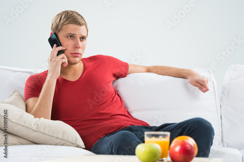 Photo: Serious young man sitting on sofa and calling with his smart pho Serious young man sitting on sofa and calling with his smart pho