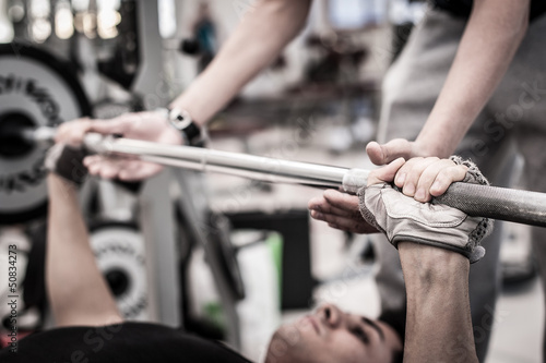 Photo: Young man lifting the barbell in the gym with instructor. Young man lifting the barbell in the gym with instructor.