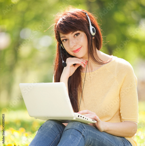 Photo: Cute woman in earphones with white laptop in the park Cute woman in earphones with white laptop in the park