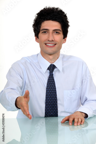 Photo: Businessman sat at desk offering to shake-hand Businessman sat at desk offering to shake-hand