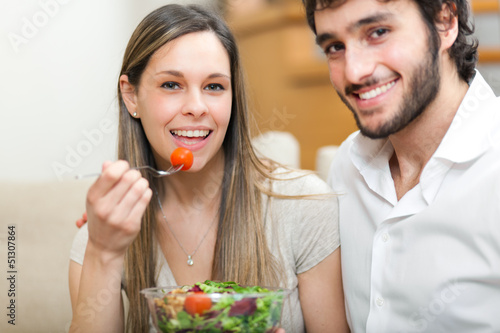 Photo: Loving happy couple eating salad in the kitchen - indoors Loving happy couple eating salad in the kitchen - indoors