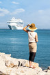 Woman with hat stands and looks at the sea, a cruise ship