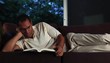 Mature man lying on sofa at home in evening reading a book