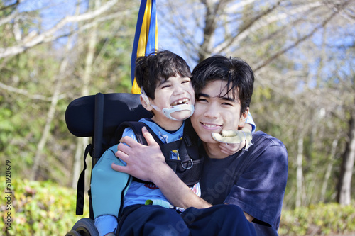 Photo: Little disabled boy in wheelchair hugging older brother outdoors Little disabled boy in wheelchair hugging older brother outdoors