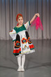 Little girl in folk costume with red wrap performs on stage.