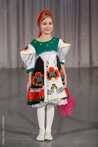 Photo: smiling little girl in folk costume with wrap performs on stage. smiling little girl in folk costume with wrap performs on stage.