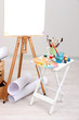 Wooden easel with clean paper and art supplies in room
