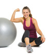 Happy woman sitting near fitness ball and showing muscles