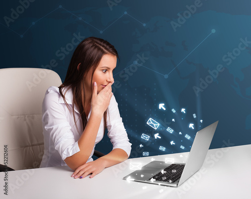 Photo: Young lady sitting at dest and typing on laptop with message ico Young lady sitting at dest and typing on laptop with message ico