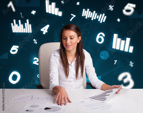 Photo: Young businesswoman sitting at desk with diagrams and statistics Young businesswoman sitting at desk with diagrams and statistics