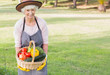Smiling elderly woman carrying basket of vegetables wearing hat and apron