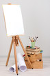 Wooden easel with clean paper and art supplies in room
