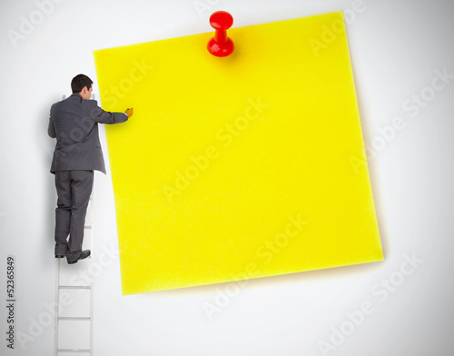 Photo: Businessman standing on ladder writing on large yellow note Businessman standing on ladder writing on large yellow note