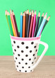 Colorful pencils in cup on table on green background