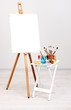 Wooden easel with clean paper and art supplies in room