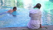 Woman with cellphone sit by the pool, man swimming