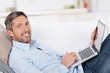 lächelnder mann liegt mit laptop auf dem sofa