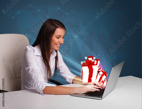 Photo: Pretty lady sitting at desk and typing on laptop with present bo Pretty lady sitting at desk and typing on laptop with present bo