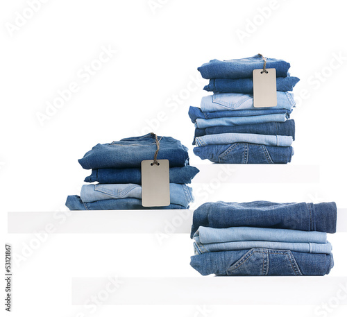 Photo: Pile of blue jeans with tag label on white shelf in shop. Pile of blue jeans with tag label on white shelf in shop.