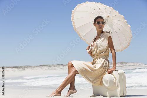 Photo: Beautiful woman sitting on a suitcase with an umbrella on the beach Beautiful woman sitting on a suitcase with an umbrella on the beach