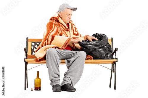 Photo: Homeless mature man sitting on a bench and looking for something Homeless mature man sitting on a bench and looking for something