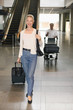 Photo: Woman pulling luggage, man pushing luggage cart in the background while text messaging on phone Woman pulling luggage, man pushing luggage cart in the background while text messaging on phone