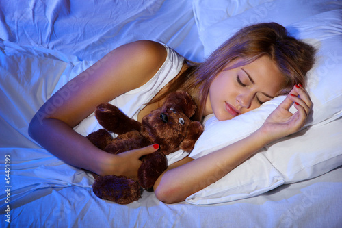 Photo: young beautiful woman with toy bear sleeping on bed in bedroom young beautiful woman with toy bear sleeping on bed in bedroom