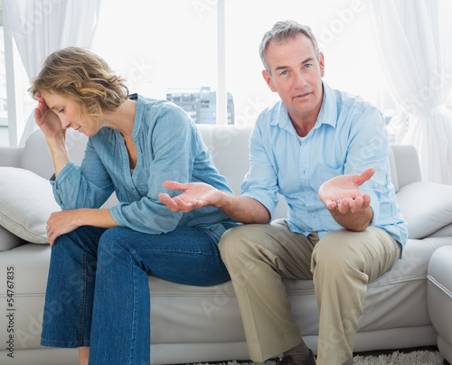Photo: Arguing middle aged couple sitting on the couch with man gesturi Arguing middle aged couple sitting on the couch with man gesturi
