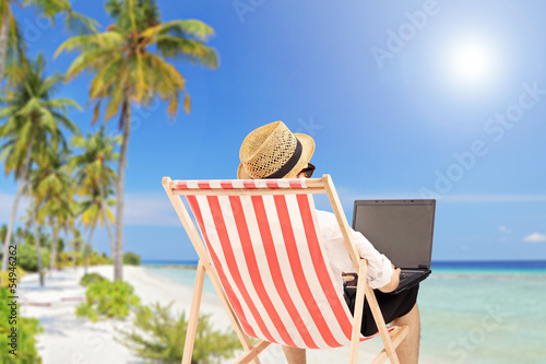Photo: Young man on an outdoor chair working on a laptop, on a beach Young man on an outdoor chair working on a laptop, on a beach