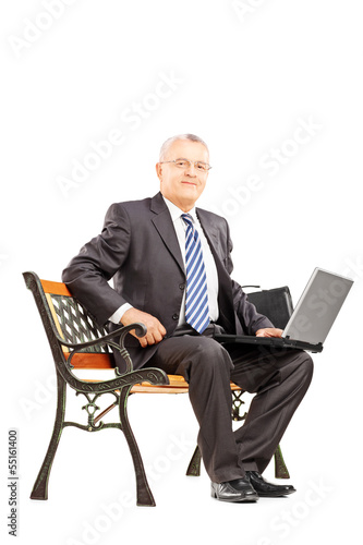 Photo: Mature businessman sitting on a wooden bench with laptop Mature businessman sitting on a wooden bench with laptop