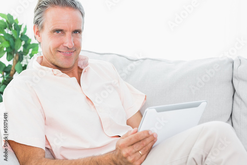 Photo: Cheerful man on his couch using tablet pc looking at camera Cheerful man on his couch using tablet pc looking at camera