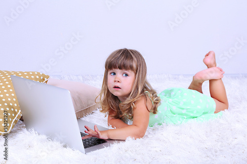 Photo: Little cute girl lies on carpet with laptop, on gray background Little cute girl lies on carpet with laptop, on gray background
