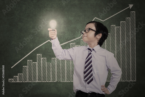 Photo: Confident business boy holding lit bulb on bar chart Confident business boy holding lit bulb on bar chart