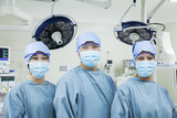 Photo: Portrait of three surgeons in a row wearing surgical masks in the operating room, looking at camera Portrait of three surgeons in a row wearing surgical masks in the operating room, looking at camera