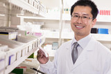 Photo: Pharmacist taking down and examining prescription medication in a pharmacy, looking at camera Pharmacist taking down and examining prescription medication in a pharmacy, looking at camera