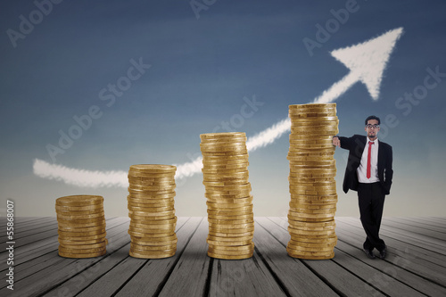 Photo: Asian businessman standing next to gold coins chart Asian businessman standing next to gold coins chart