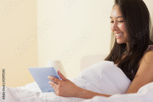 Photo: Happy young asian woman sitting in bed using tablet pc Happy young asian woman sitting in bed using tablet pc