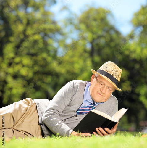 Photo: Senior man with hat lying on a grass reading a book in a park Senior man with hat lying on a grass reading a book in a park